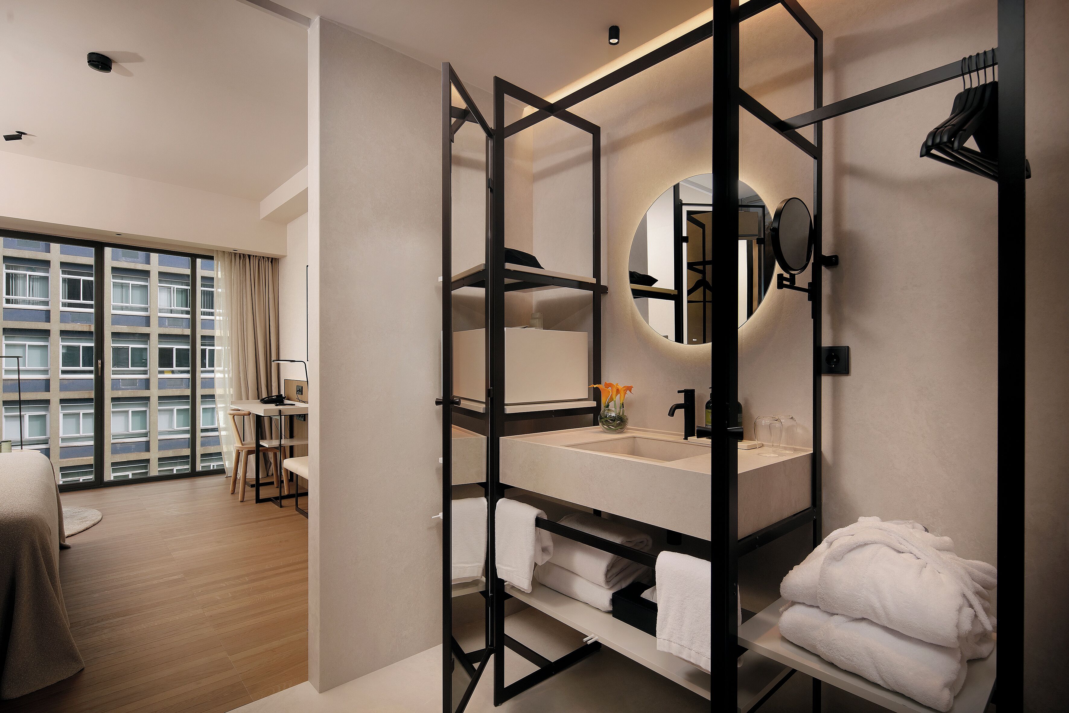 A modern hotel room with a large window, a separate sitting area, a bathroom with a vanity, and a mirror.