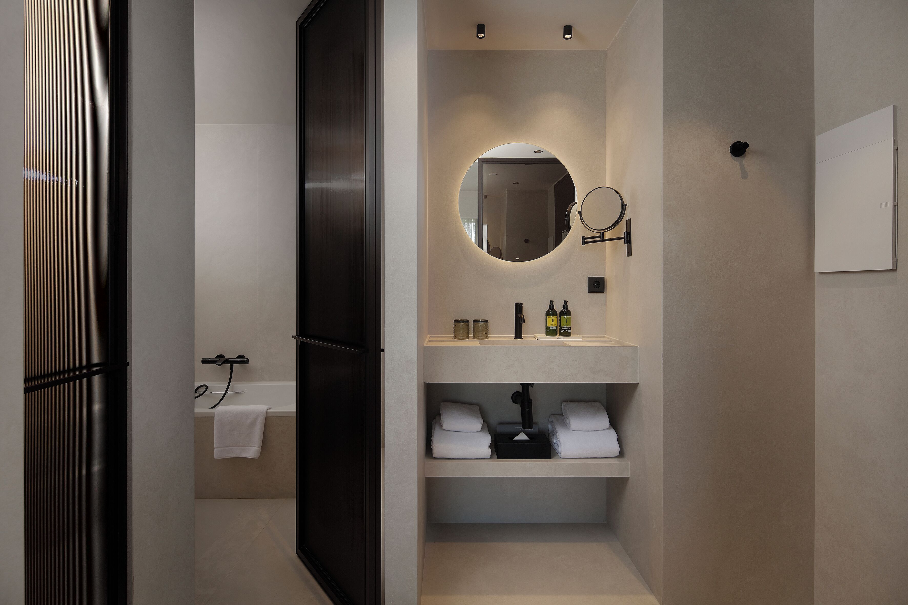 A modern bathroom with a bathtub, a round mirror, and shelves with towels and toiletries.