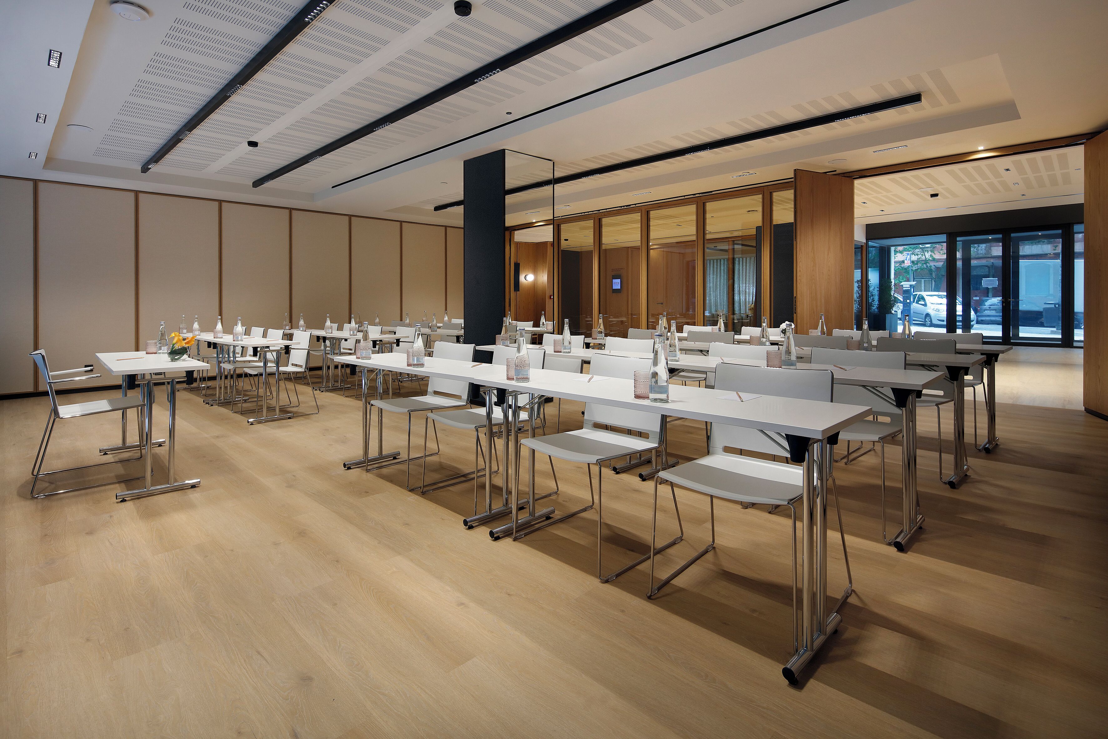 Un salón de conferencias con mesas y sillas blancas, suelo de madera y ventanas amplias con vistas al exterior.