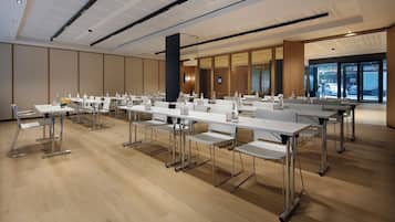 Un salón de conferencias con mesas y sillas blancas, suelo de madera y ventanas amplias con vistas al exterior.