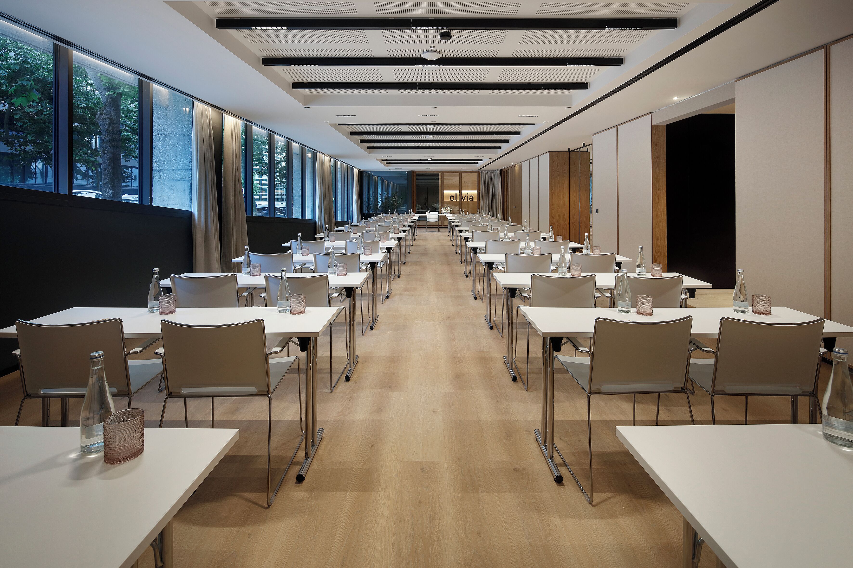Sala de conferências com mesas compridas, cadeiras e vista para árvores através de amplas janelas.