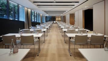 Sala de conferências com mesas compridas, cadeiras e vista para árvores através de amplas janelas.