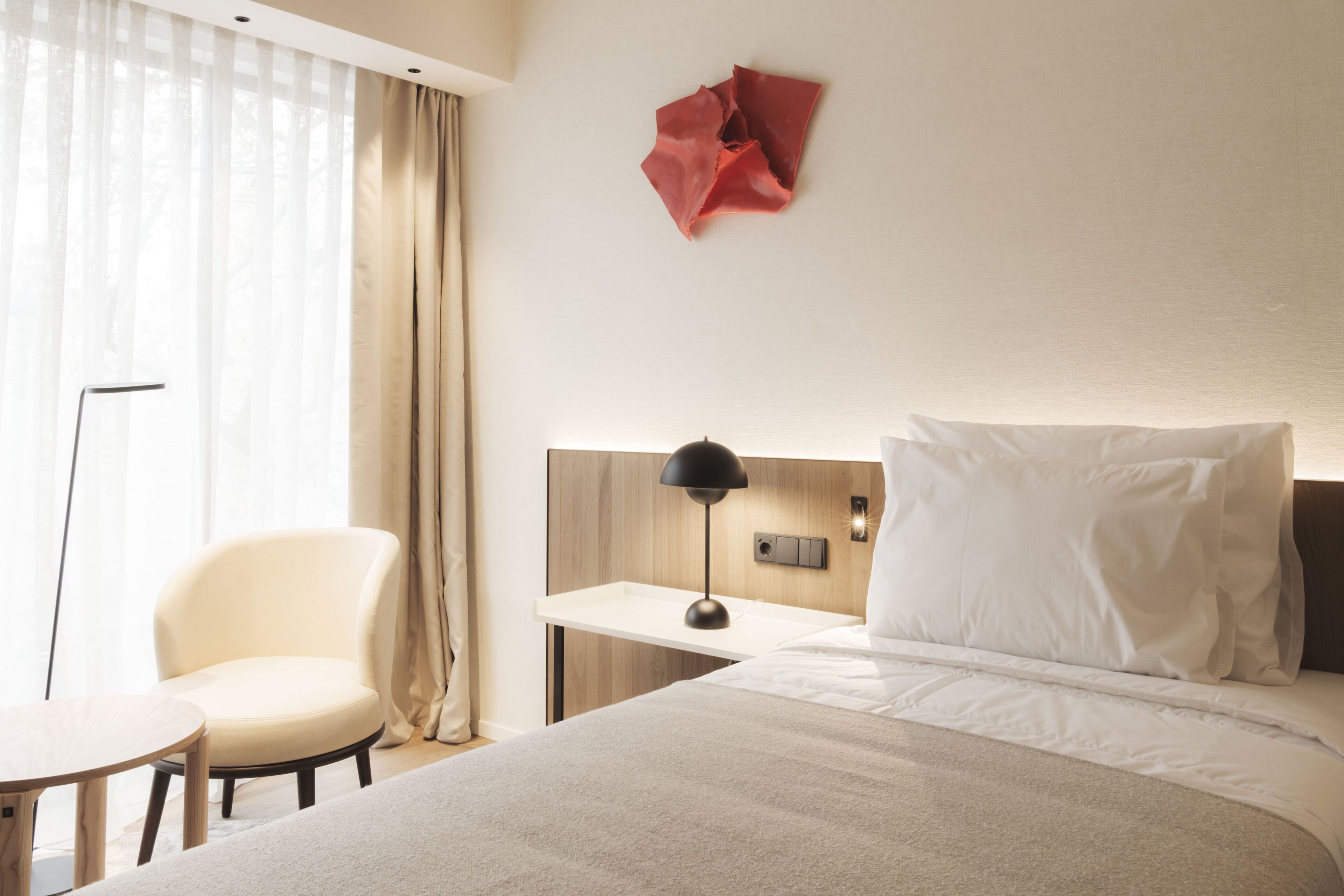A hotel room with a bed, a bedside table, a lamp, a chair, and a window with curtains.