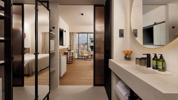 Habitación de hotel con cama, escritorio, silla, zona de comedor y baño con un espejo grande.