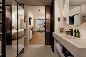 Habitación de hotel con cama, escritorio, silla, zona de comedor y baño con un espejo grande.