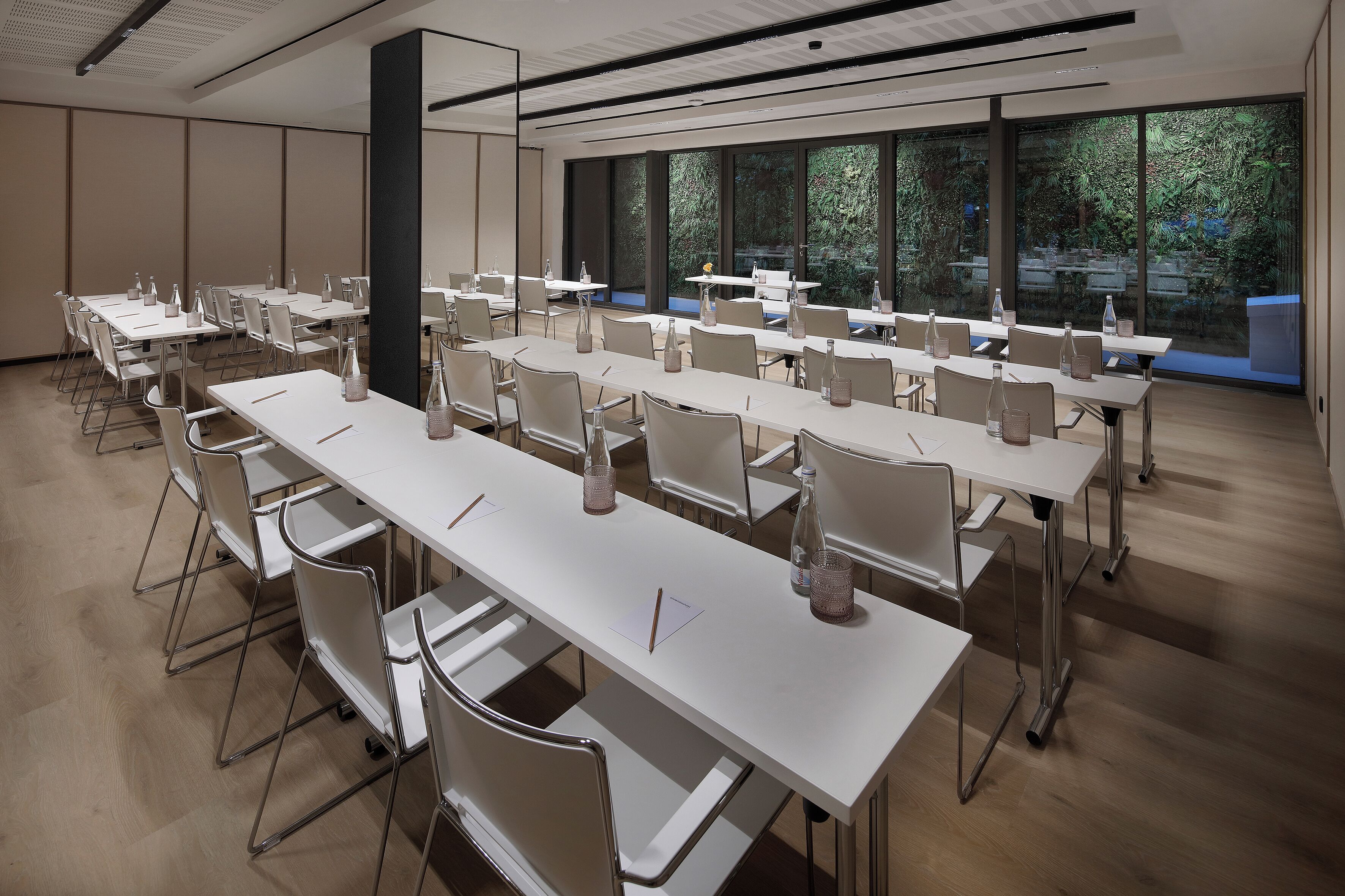 A conference room with multiple tables and chairs, large windows with a view of greenery, and a modern ceiling design.