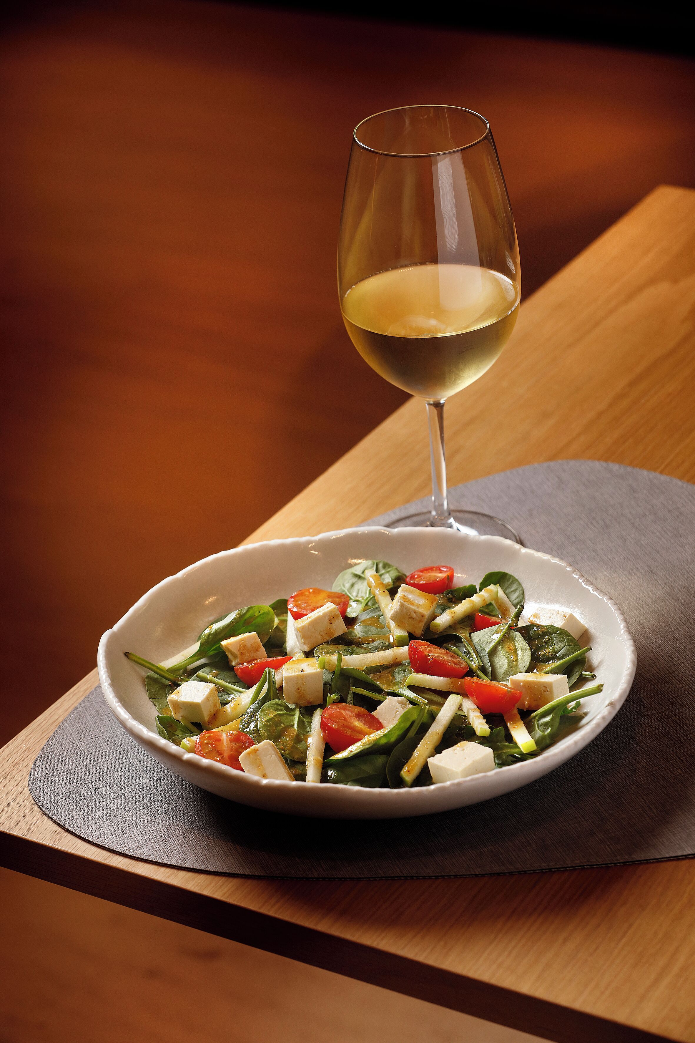 A white bowl of salad with a glass of white wine on a wooden table.