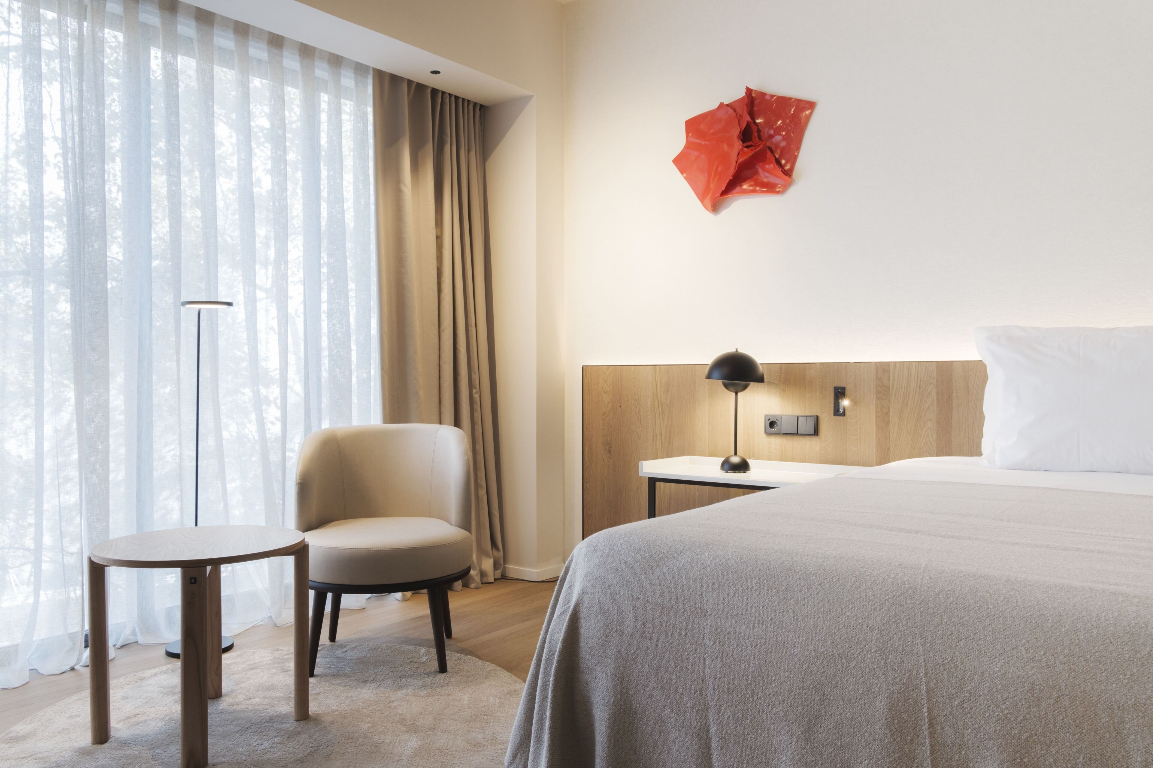 A hotel room with a bed, a chair, a small table, a lamp, and a red abstract art piece on the wall.