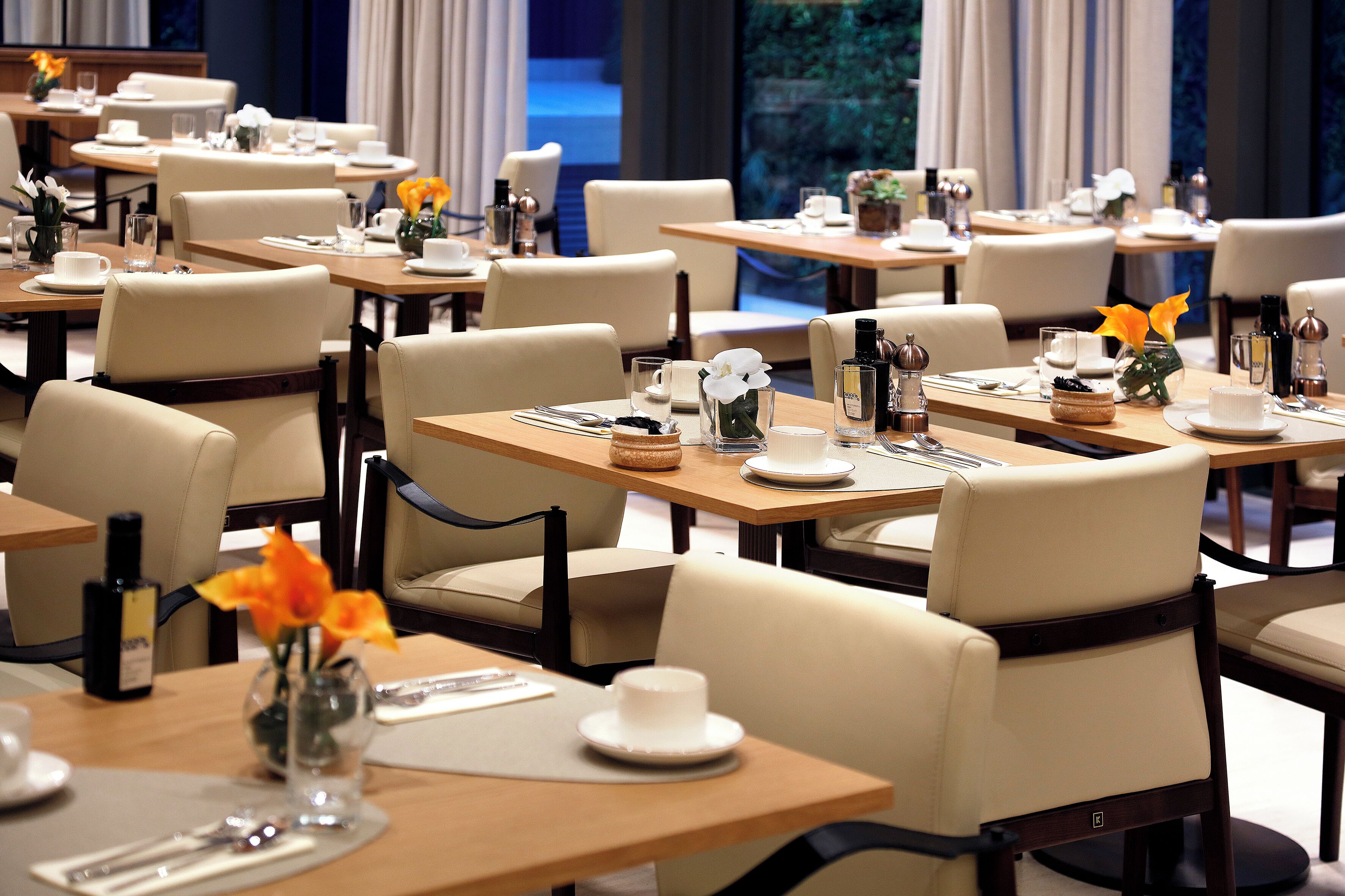 A hotel dining area with multiple tables, chairs, and neatly set dining tables with plates, glasses, and cutlery.