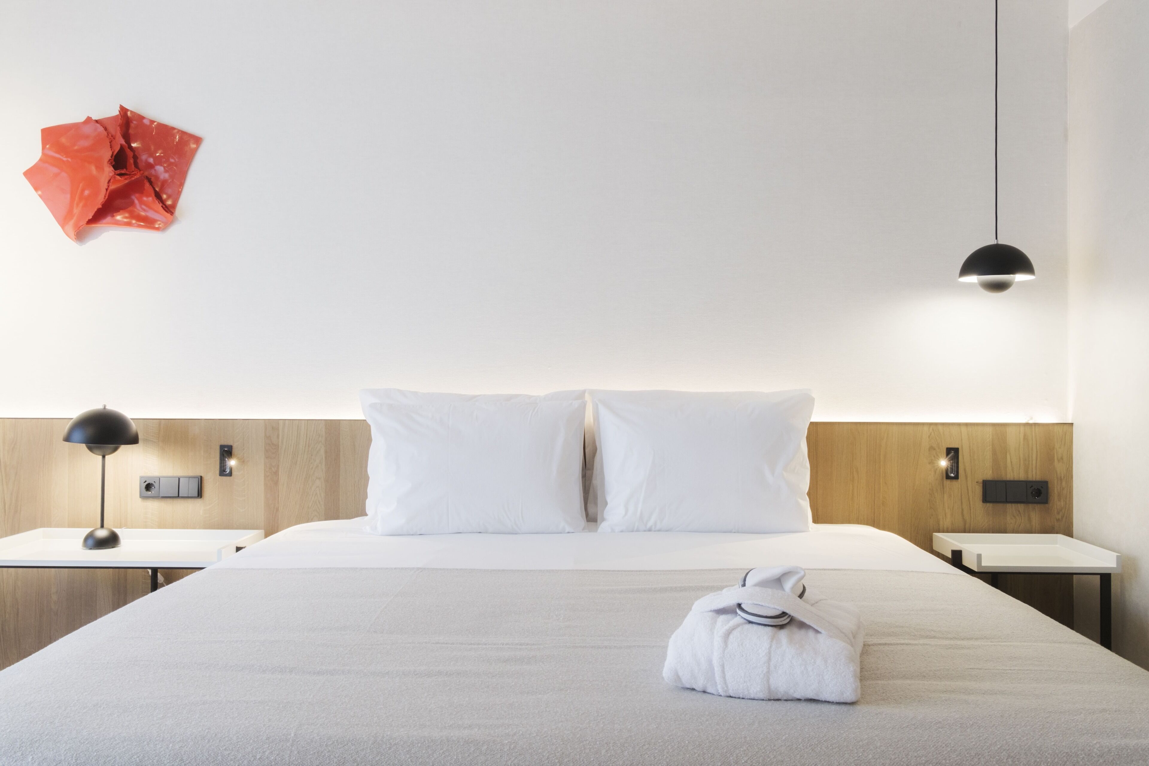 A hotel room with a bed, two bedside tables, a lamp, a wall-mounted light, and a red decorative item.