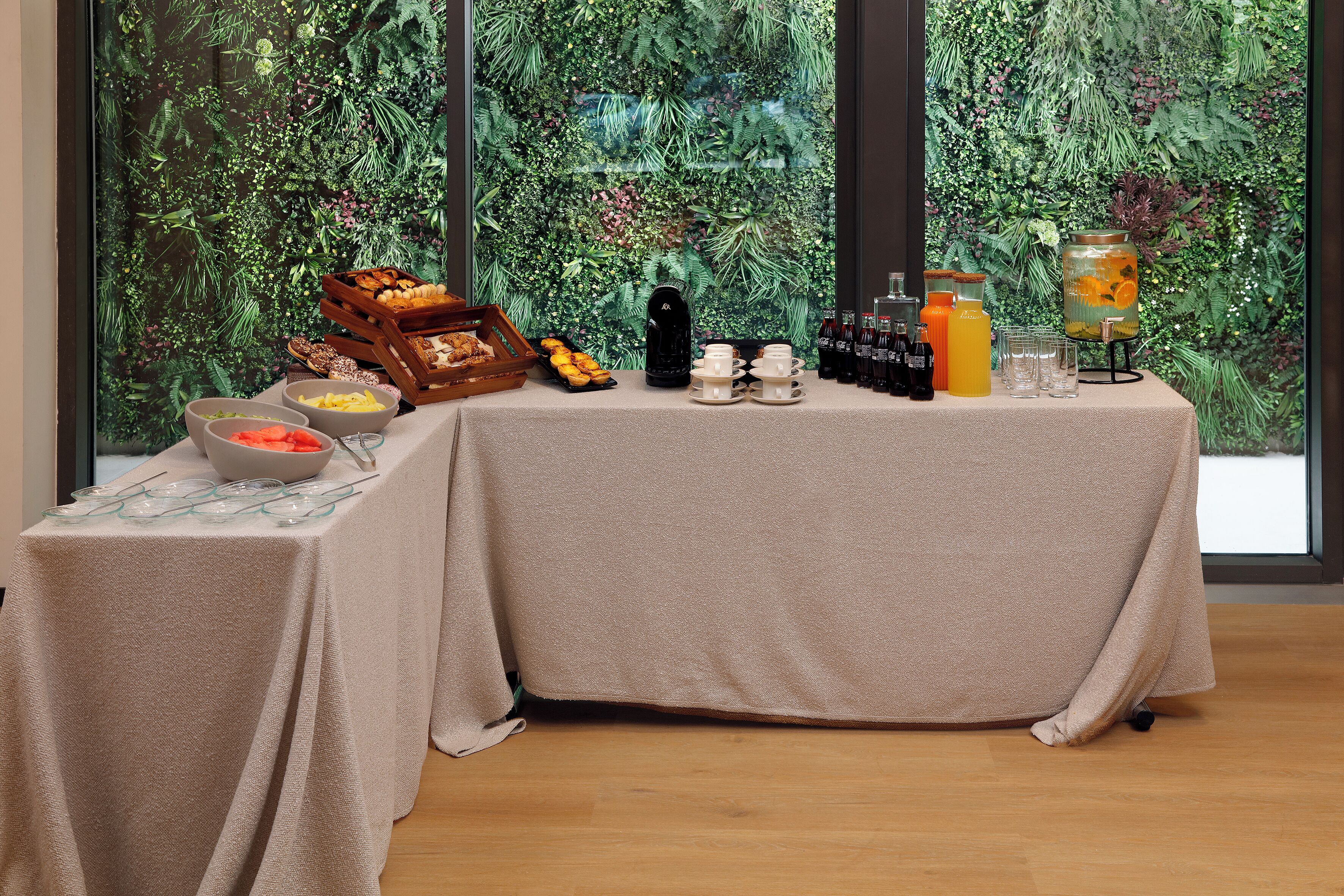 A table set with breakfast items, a fruit bowl, and a beverage dispenser, with a lush green backdrop visible through the window.