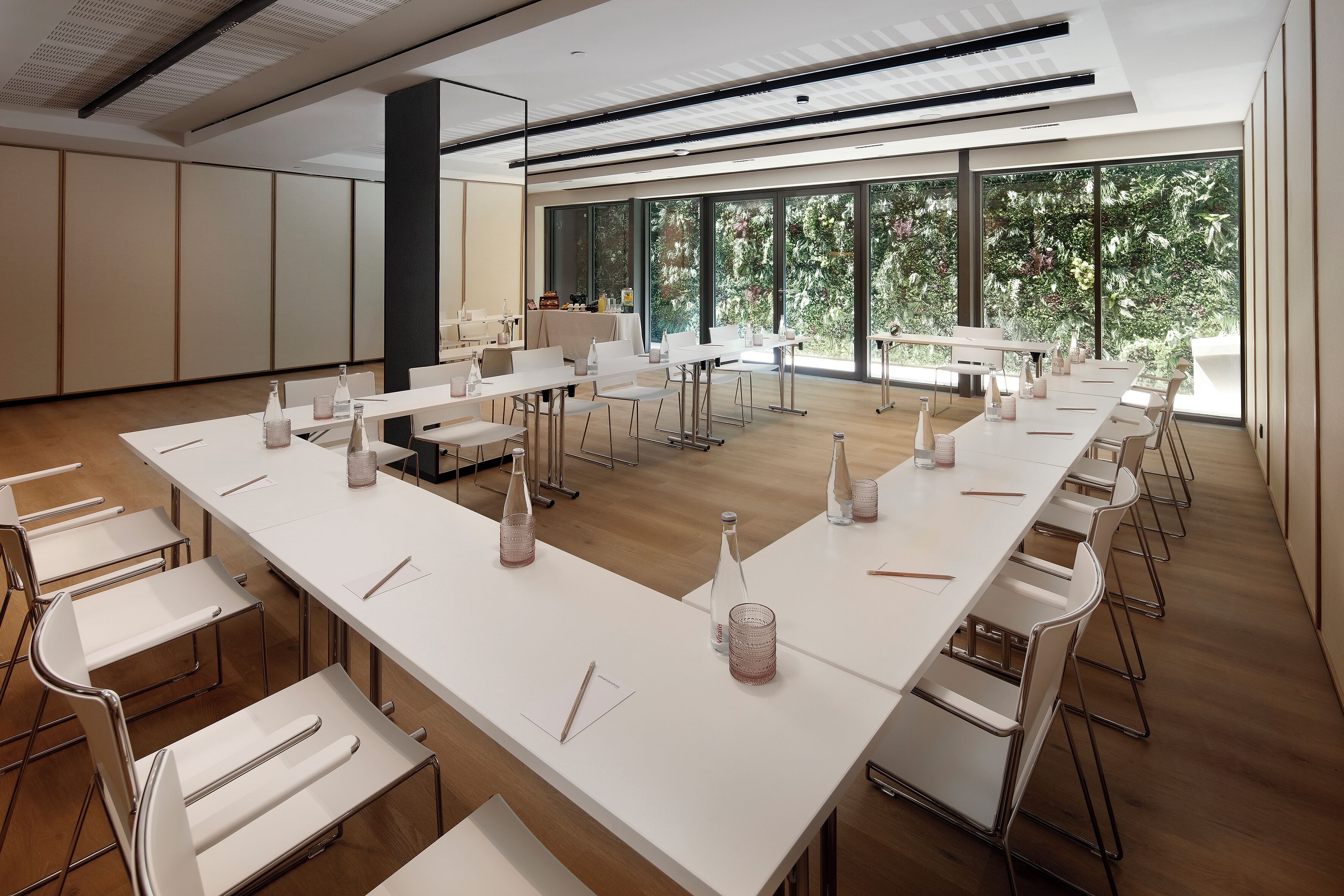 Une salle de conférence dotée de longues tables, de chaises et de grandes fenêtres donnant sur un paysage arboré.