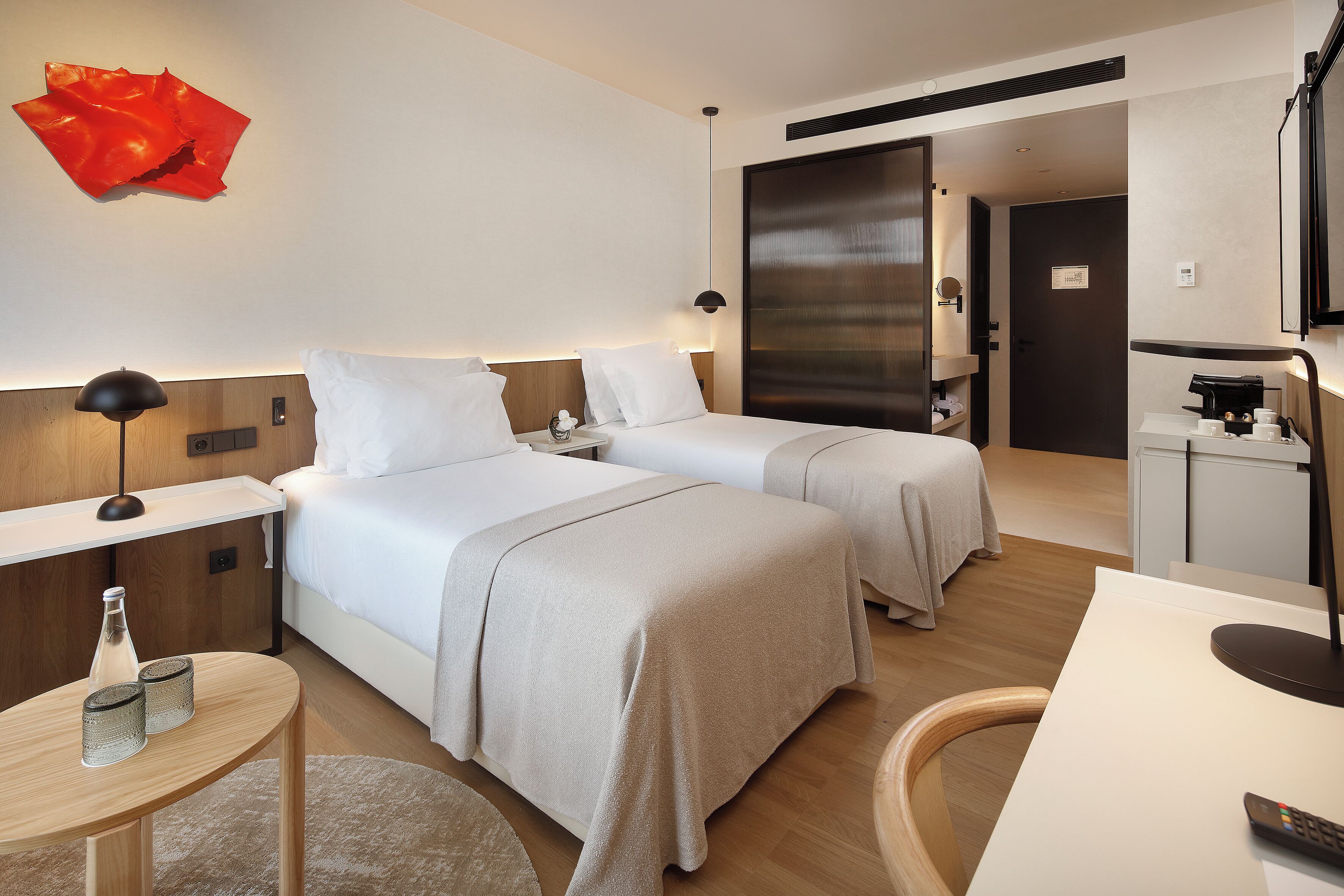 Habitación de hotel con dos camas, un escritorio, una silla, una lámpara, una cafetera y una obra de arte abstracta roja en la pared.