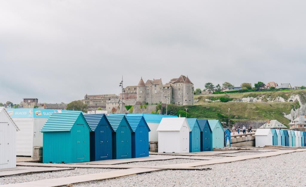 Een kustgebied met een kasteel op de achtergrond en een rij blauw-witte strandhutjes op de voorgrond.