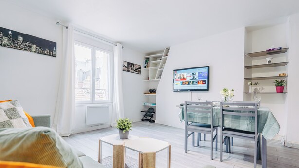 Moderni olohuone, jossa on sohva, ruokapöytä tuoleineen, televisio ja seinällä kaupunkimaisemaa esittävä taideteos.
