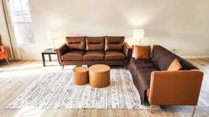 A living room with a brown leather sofa, two wooden side tables, a patterned rug, and a window with curtains. - Near ​JT Park: Hot Tub, Cinema, Arcade Game, GameRM  (Yucca Valley)