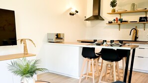 A modern kitchen with a central island, a dining area with two chairs, a sofa with a colorful cushion, and a TV mounted on the wall. - Apartment 5 min from Disney (Montévrain)