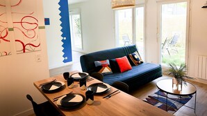 A modern living room with a dining table set for four, a blue sofa with colorful cushions, and a hanging rattan light fixture. - Apartment 5 min from Disney (Montévrain)