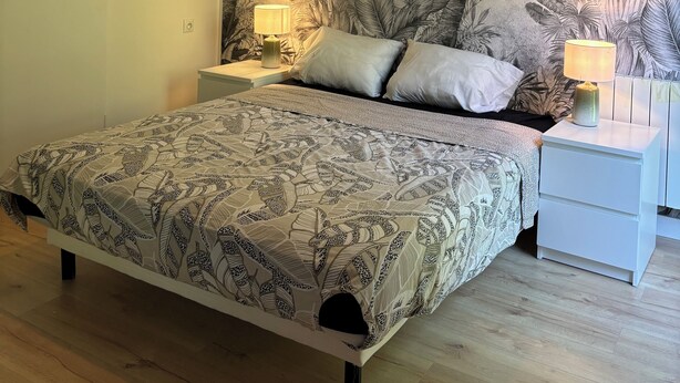 A bedroom with a bed, nightstands, a wall mural of palm trees, and a hanging lamp. - Apartment 5 min from Disney (Montévrain)