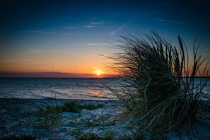 A beach scene with the sun setting over the ocean, featuring sand and grasses in the foreground. - Haus am Fjord - Private Parking Space, Washing Machine, EV Charging, Toys (Olpenitz)