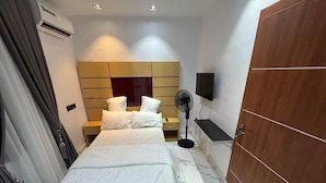 Une chambre d’hôtel avec un lit, une armoire en bois, un téléviseur fixé au mur et un ventilateur sur pied.