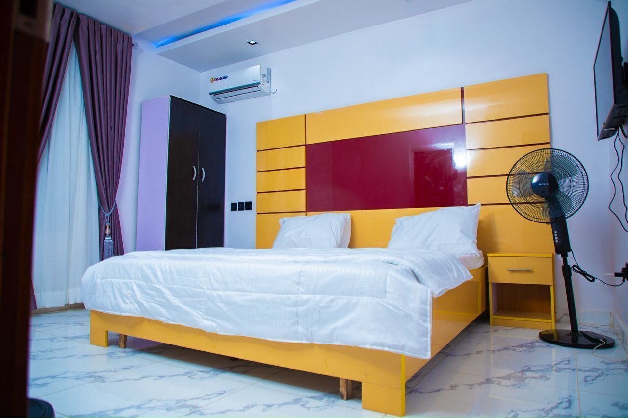 A bedroom with a bed, a bedside table, a nightstand, an air conditioner, a wall-mounted TV, and a standing fan.