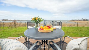 A round table with a fruit bowl and a vase of flowers, overlooking a grassy area and a body of water. - Albatros - Sea view, infrared cabin, beach chair, private parking, terrace (Olpenitz)
