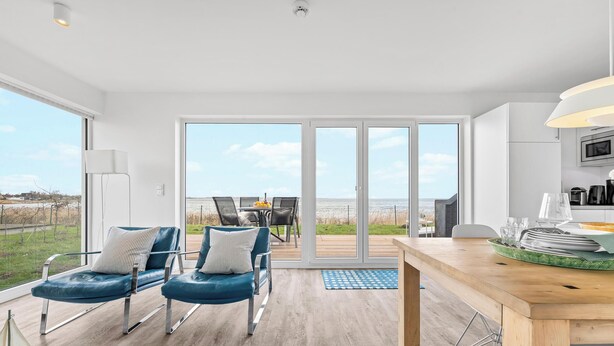A modern kitchen with a wooden dining table, a kitchen island, and a view of the sea through large sliding doors. - Albatros - Sea view, infrared cabin, beach chair, private parking, terrace (Olpenitz)