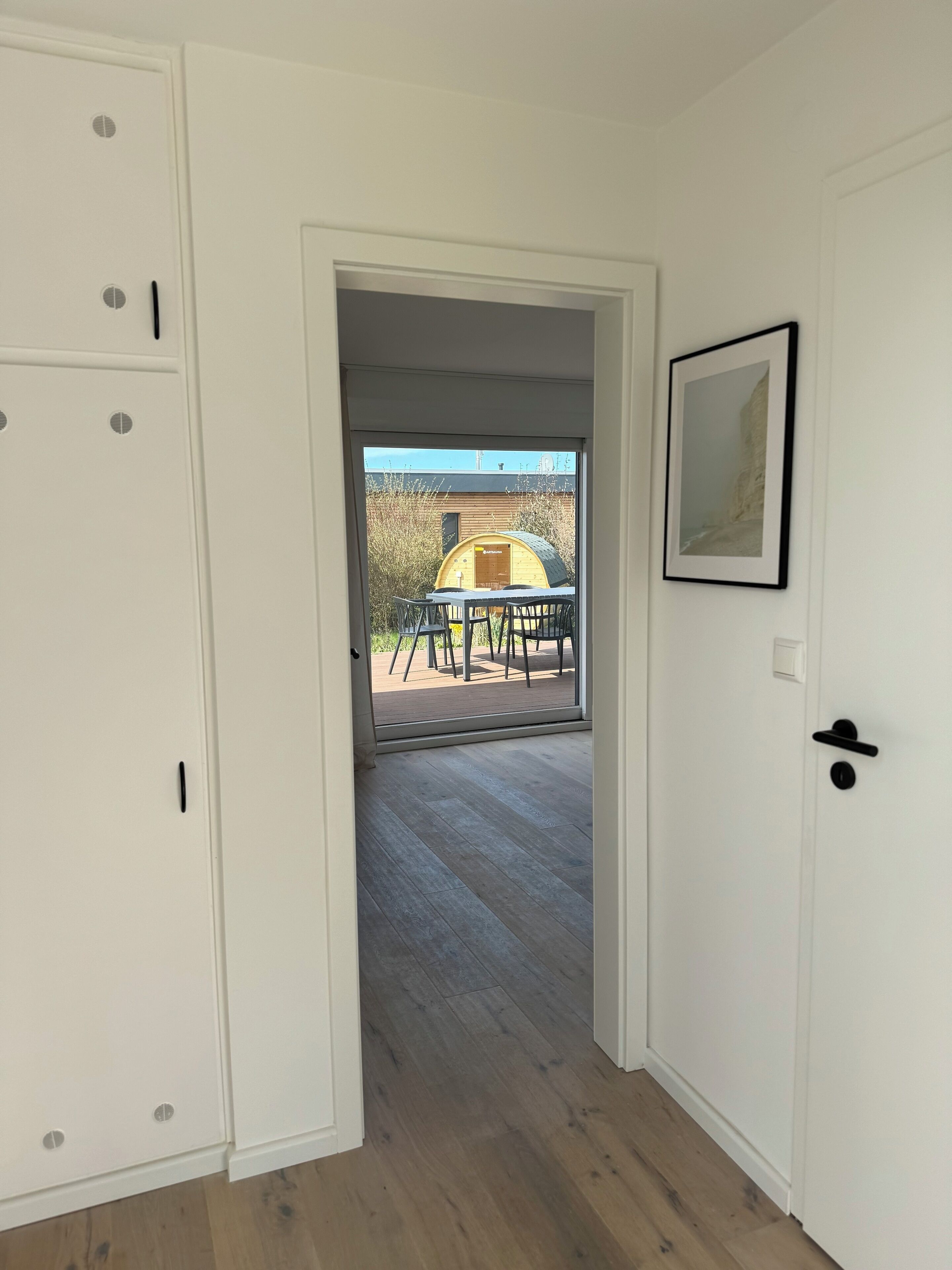 A room with a wooden floor, a white door, and a view of an outdoor area with a table and chairs.
