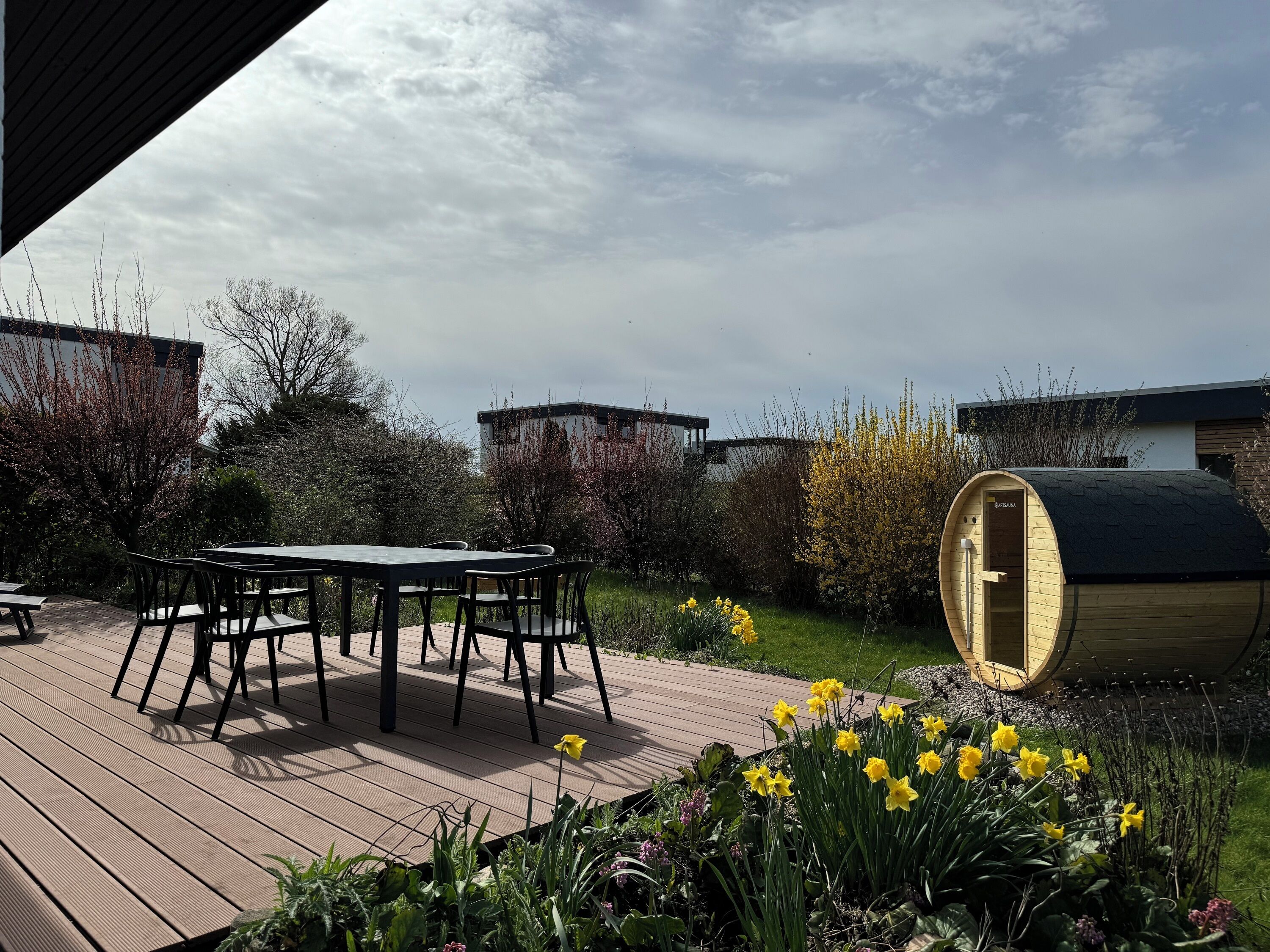 En træterrasse med et bord og stole, omgivet af en have med påskeliljer og andre planter.