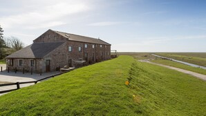 A large stone building with a sloped roof, a paved area in front, and a grassy field with a water channel.
