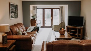 A living room with a brown leather sofa, a television on a wooden stand, a lamp, and a view through a glass door to the outside. - The Lucky Duck, Patty's Barn, Lancaster, Cockerham (Lancaster)