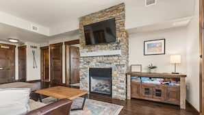 A living room with a stone fireplace, a flat-screen TV mounted on the wall, a wooden coffee table, a leather sofa, and a bookshelf with books. - 500 ft from Pickleball | Private Gondola Access | Hot Tubs & Fitness Center (Steamboat Springs)