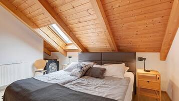 Ein Schlafzimmer mit schrägverlaufender Holzdachkonstruktion, einem Bett mit Kissen, einem Nachttisch mit Lampe und einem Heizkörper.