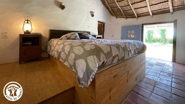 A rustic bedroom with a thatched roof, a wooden bed, a nightstand with a lantern, and a view of greenery outside.