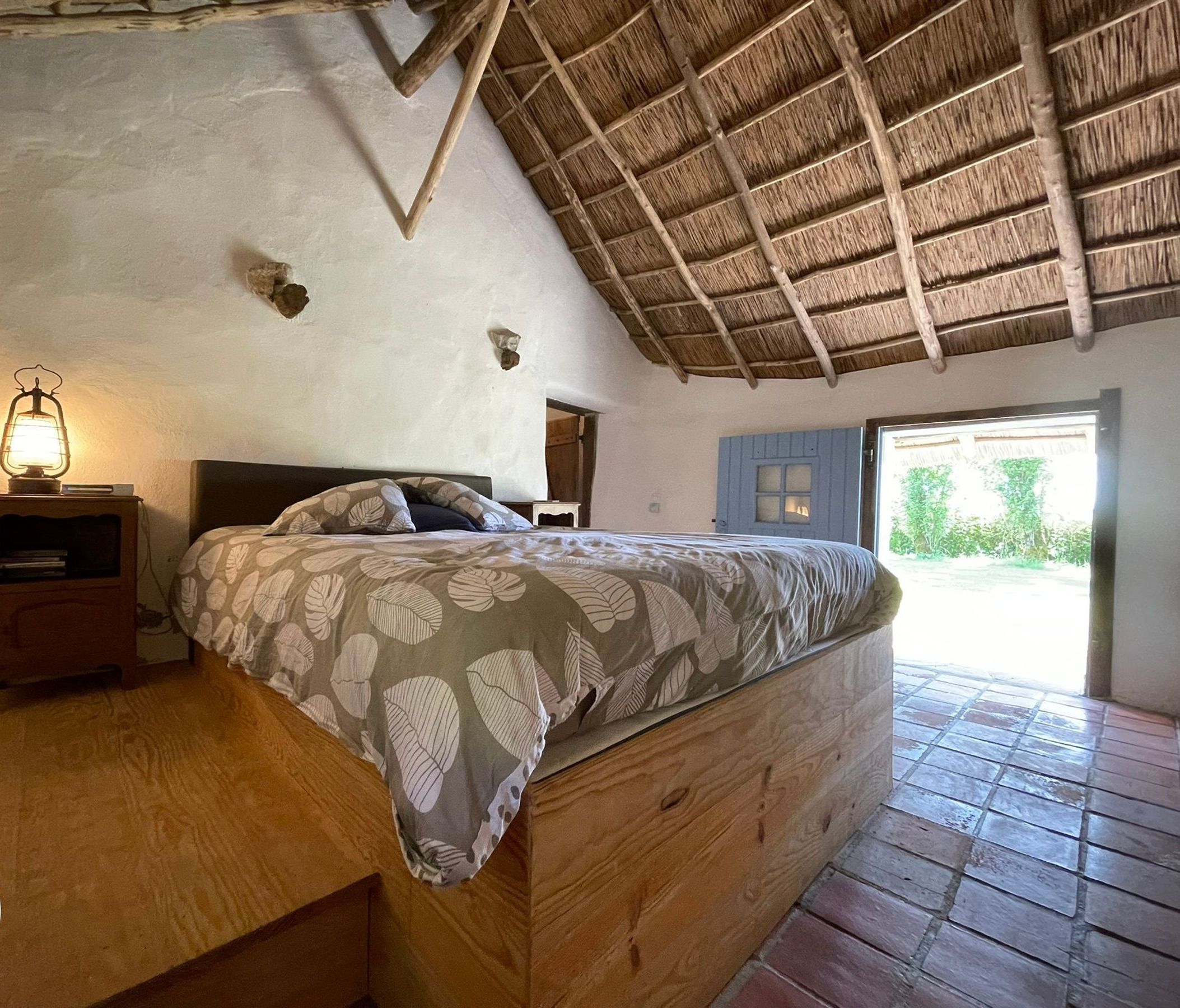 Une chambre rustique avec un toit de chaume, un lit en bois, une table de chevet avec une lanterne et une vue sur la verdure environnante.