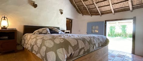 A rustic bedroom with a thatched roof, a wooden bed, a nightstand with a lantern, and a view of greenery outside.