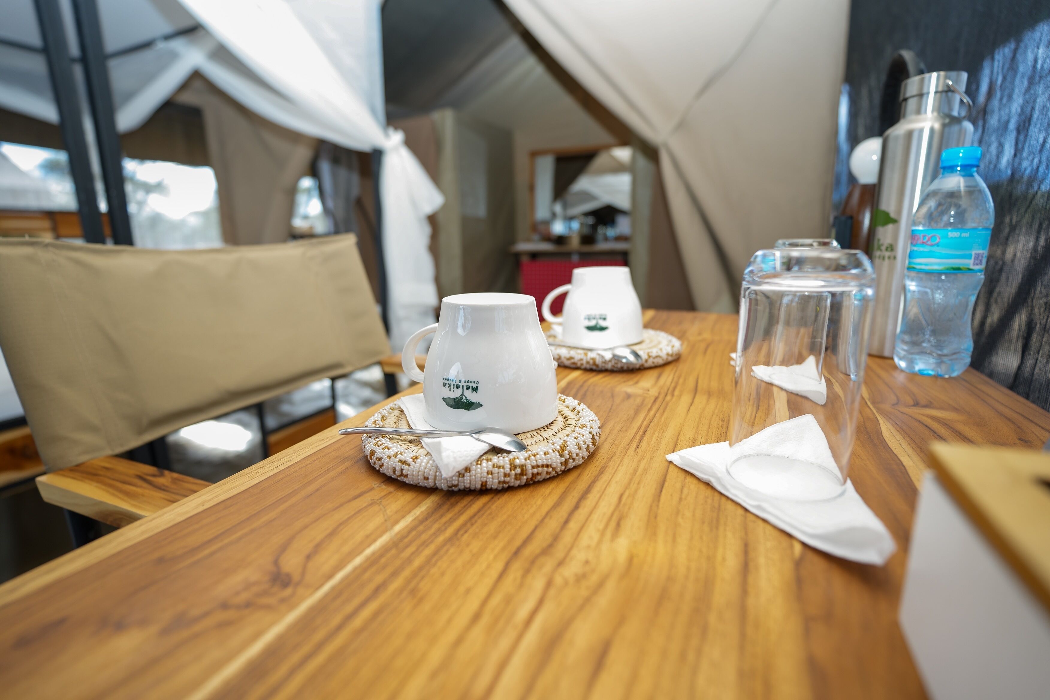 A wooden table with two white cups and saucers, a water bottle, and a thermos.