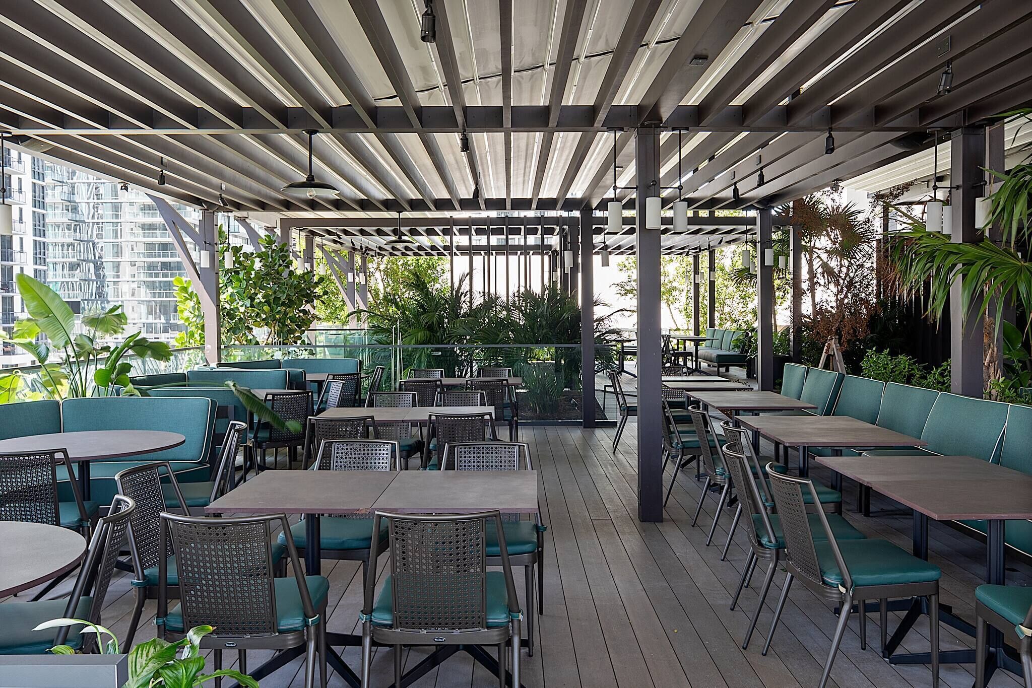 An outdoor patio with tables and chairs, surrounded by greenery and a modern building in the background.