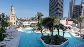 Ein Pool auf dem Dach mit Palmen, Liegestühlen und Blick auf die Stadtlandschaft, darunter ein historischer Turm und moderne Gebäude.
