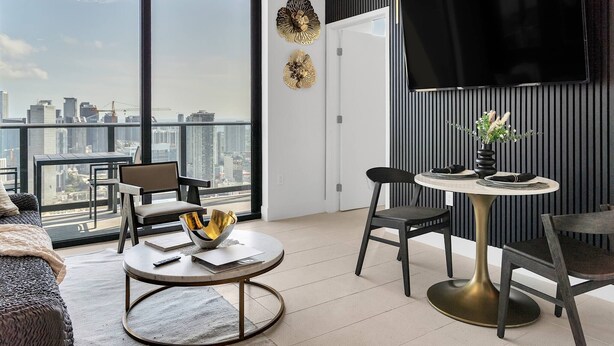A modern living room with a city view, a round table, two chairs, a sofa, and a TV mounted on the wall.