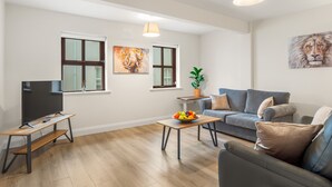 A modern living room with a grey sofa, a wooden coffee table with fruit, a TV on a stand, and artwork on the walls. - 17 Clifden Court getaway with free parking (Clifden)