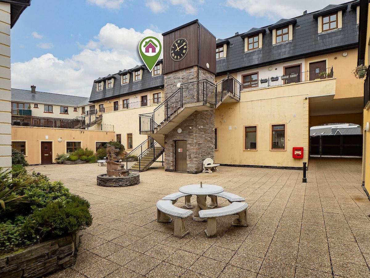 A courtyard with a stone bench, a clock tower, and a staircase leading to an upper level.