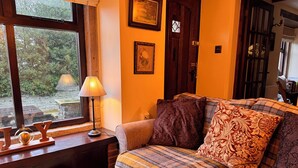 Ein gemütliches Zimmer mit Holzboden, einer Couch mit gemusterten Kissen, einem kleinen Tisch mit Lampe und einem Fenster mit Blick auf Grünflächen.