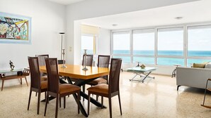 Un comedor moderno con una mesa y sillas de madera, una mesa de centro de vidrio y vistas al océano a través de amplios ventanales.
