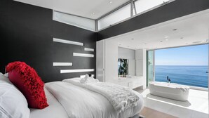 Una camera da letto moderna con un letto grande, un cuscino rosso, una coperta bianca, una vasca da bagno e vista sul mare.