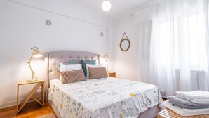Une chambre à coucher bien rangée, avec un lit, deux lampes de chevet, une table de chevet, un miroir rond et une fenêtre ornée de rideaux légers.