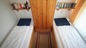Quarto pequeno com duas camas de solteiro, guarda-roupa de madeira e estantes com livros e uma miniatura de farol.