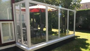 A glass-enclosed patio area with a table and chairs, a red umbrella, and a view of the surrounding garden.