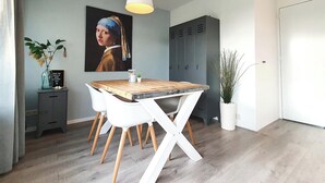 A modern dining area with a wooden table and white chairs, a pendant light, a painting on the wall, and a potted plant.